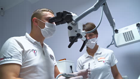 male dentist in mask and gloves stands in front of microscope. doctor is holding tools in both hands ready to apply them. low angle view.