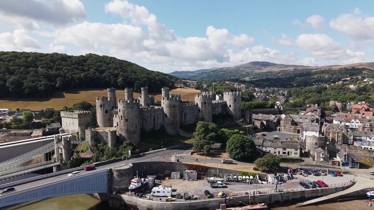Historic stone castle built by Edward I, seen from above with surrounding town, road bridge, harbour, and green hills in daylight