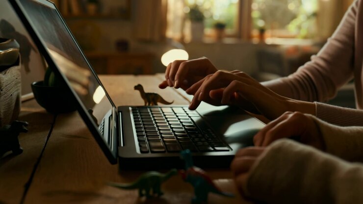 Close side view shows fingers lifting and pressing laptop keys while colorful dinosaur toys stand near the computer, with a glowing round lamp and potted plants softly blurred in the background.