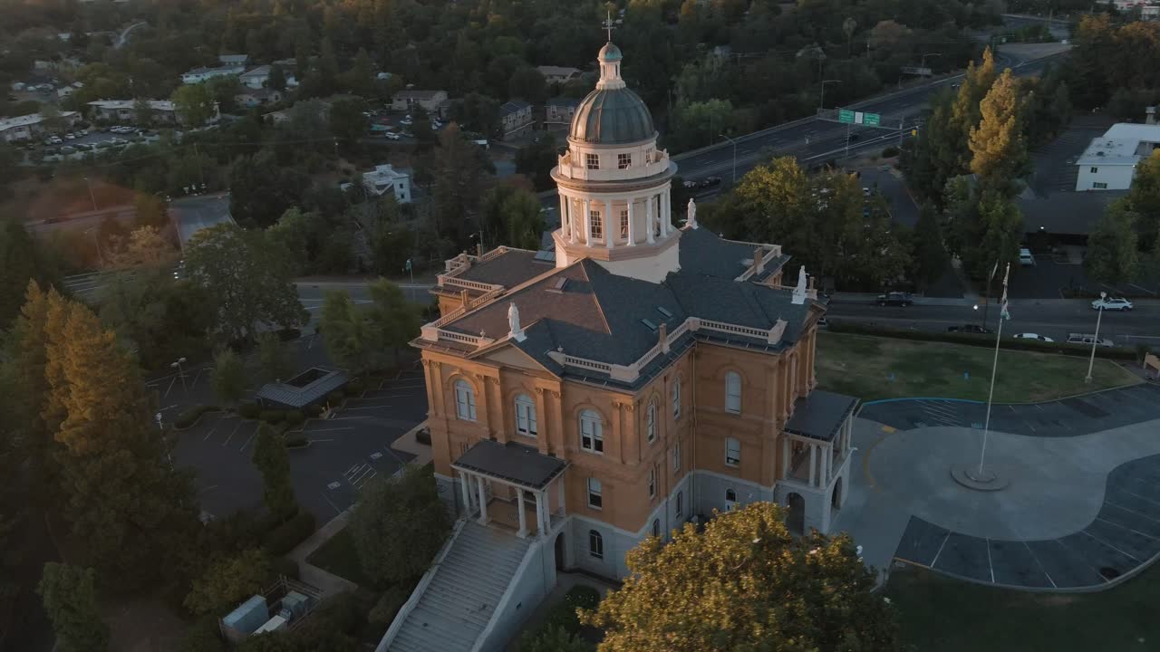 Drone orbits Auburn Courthouse, showing flag, plaza, parking, highway, trees, and nearby houses at sunset