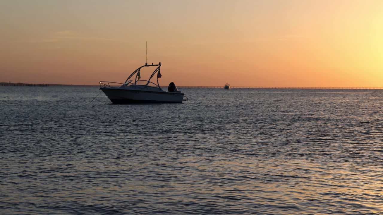 Slow motion view of small boat anchored just offshore as sun sets in background with golden sky. Chesapeake Bay Bridge Tunnel at sunset from First Landing. Glow of evening light on calm water surface.