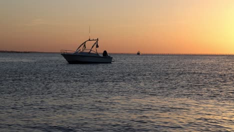 slow motion view of small boat anchored just offshore as sun sets in background with golden sky. chesapeake bay bridge tunnel at sunset from first landing. glow of evening light on calm water surface.