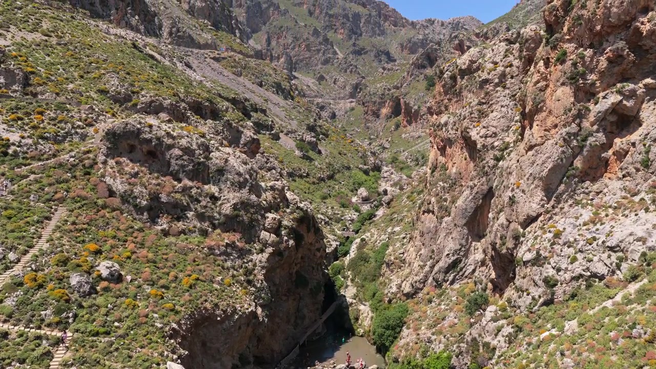Vista aerea con drone di un canyon roccioso con scogliere a picco e una piccola pozza fluviale dove le persone si radunano in fondo, Gola di Kourtaliotiko, Grecia