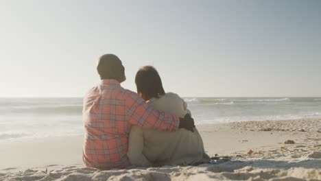 Smiling-senior-african-american-couple-embracing-and-sitting-on-sunny-beach