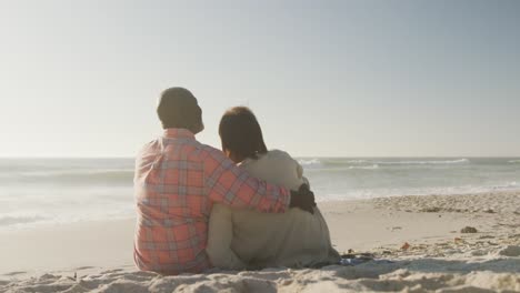 una pareja afroamericana de alto nivel sonriente abrazándose y sentada en una playa soleada