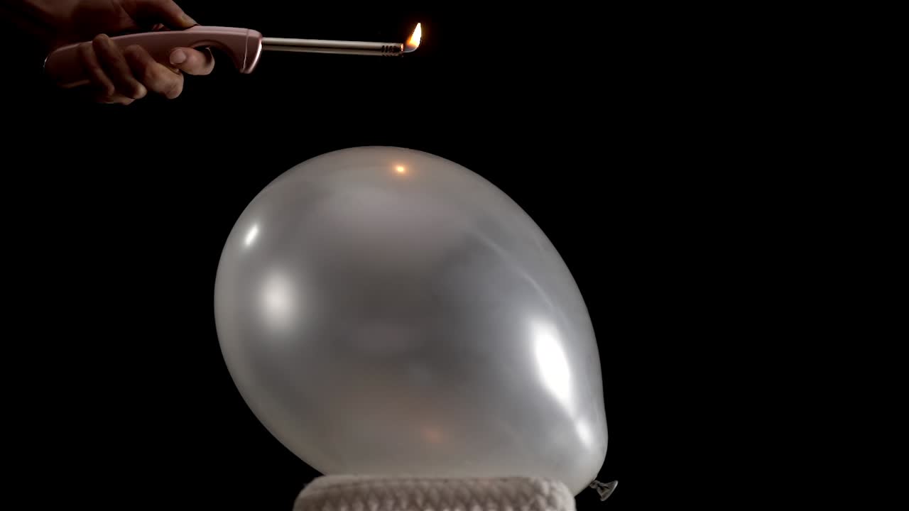 A silver balloon rests still as a hand lights a small flame on a lighter and slowly moves it closer. The tension builds until the balloon pops dramatically, captured in stunning slow motion.