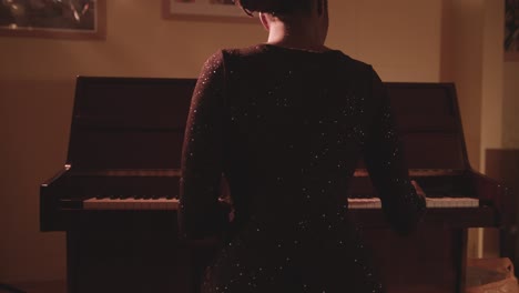 female pianist in glittery dress sits in front of a piano and plays - back view, medium shot