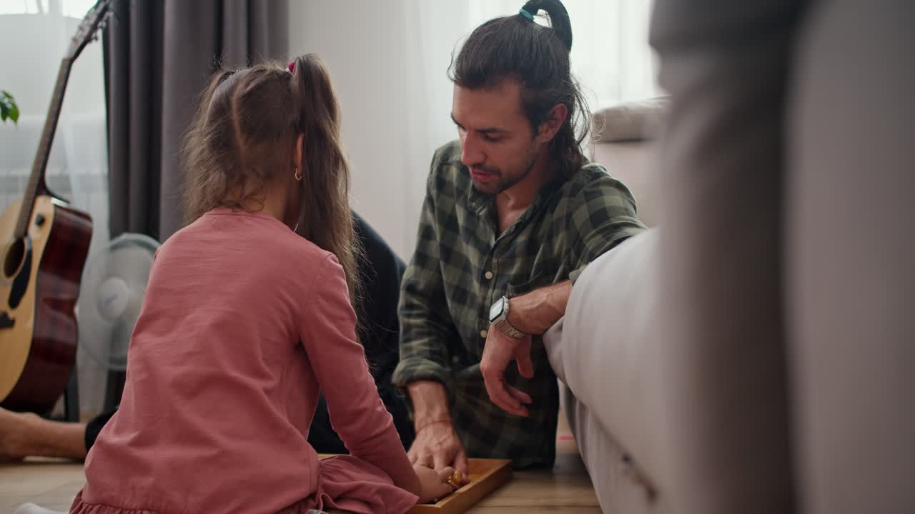 vista trasera de una pequeña chica morena en un vestido rosa juega al ajedrez con su padre, un hombre moreno en una camisa verde a cuadros, y luego ponen el juego en una caja especial apoyándose en un sofá gris en el suelo en un apartamento moderno
