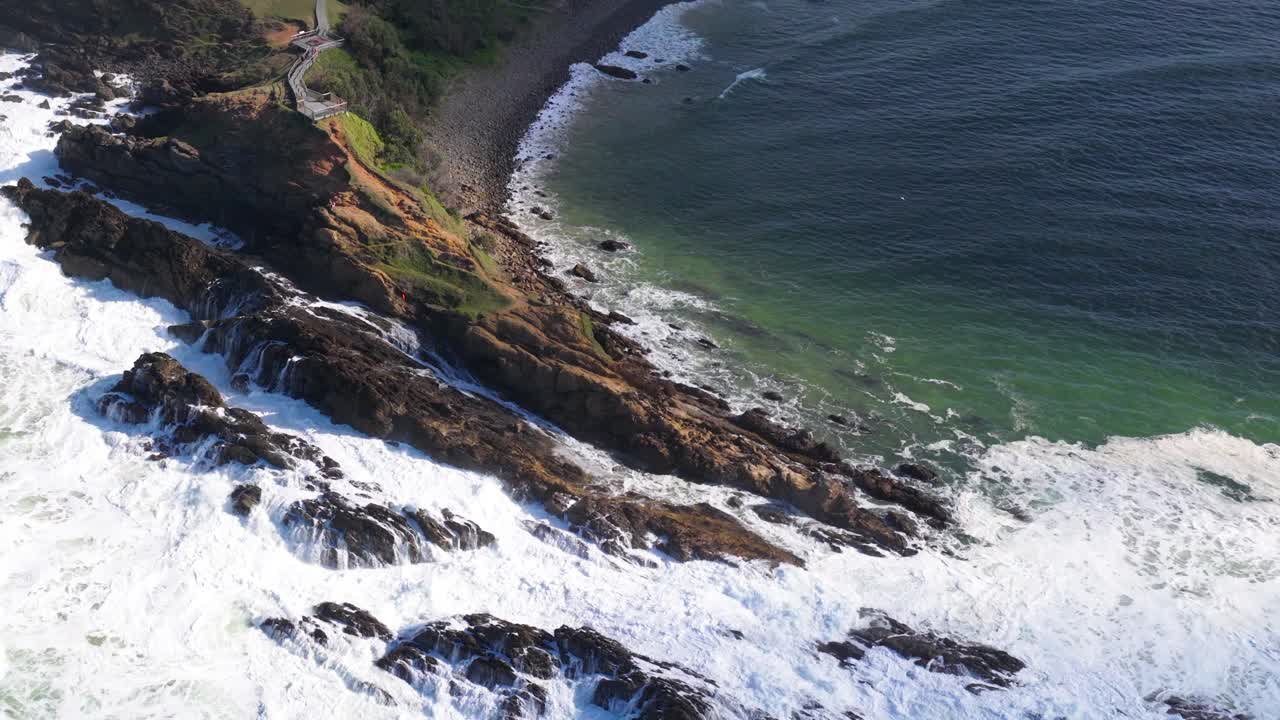 Aerial footage captures waves crashing against rocky cliffs at Byron Bay, Australia. Bright daylight enhances the vibrant coastal scenery