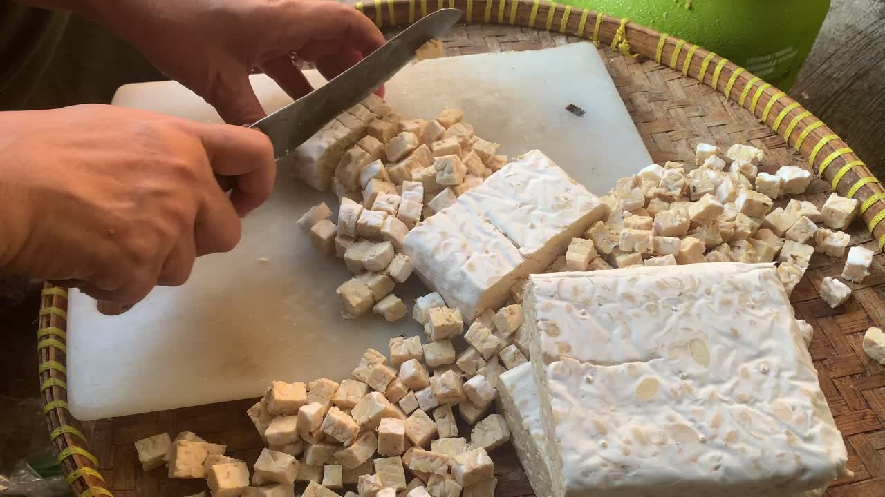 Close-up of hands cutting tempeh into small cubes on a cutting board, traditional Indonesian food preparation scene