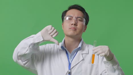 연구실에서 녹색 스크린 배경에 서있는 동안 검사 튜브에서 오렌지 액체를보고 엄지손가락을 아래로 표시하는 아시아 남성 과학자의 클로즈업