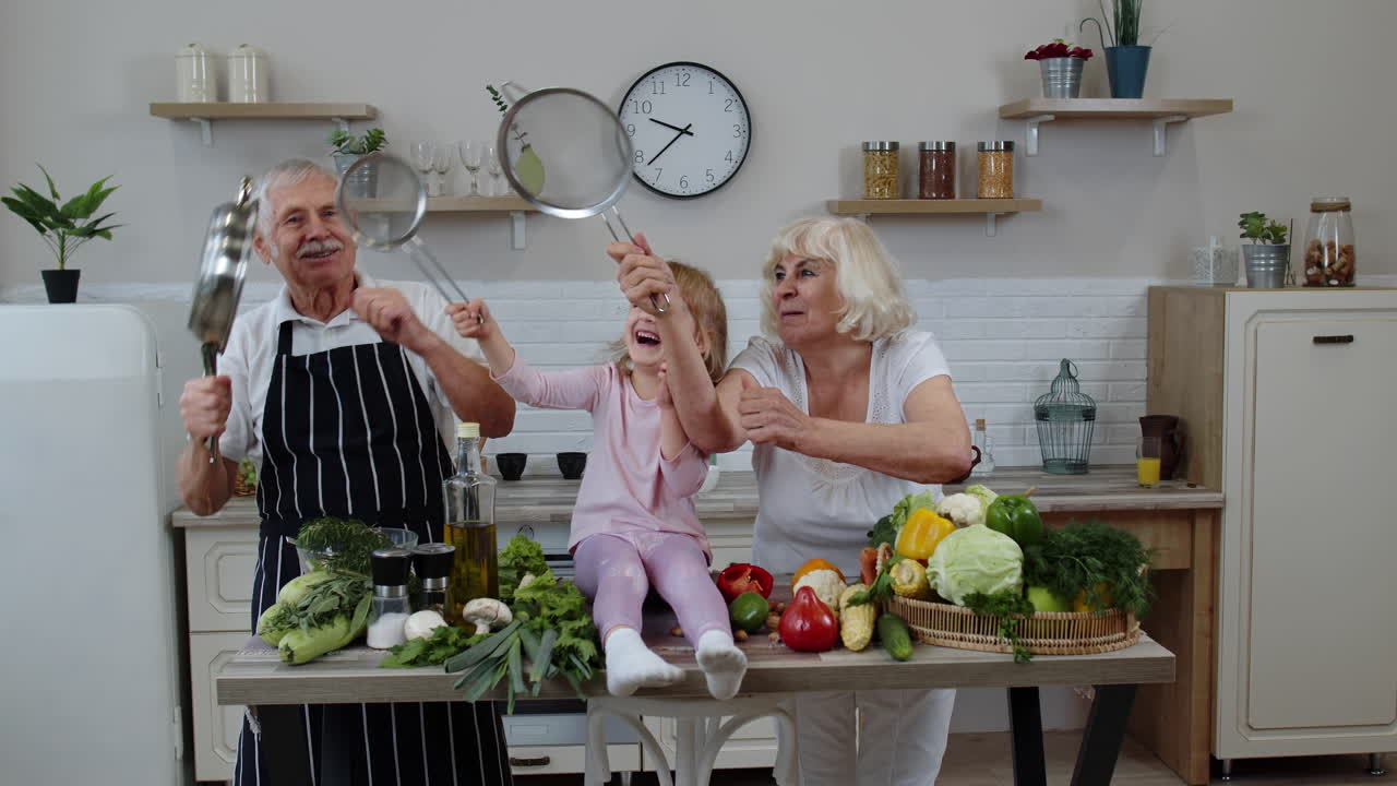 Ein glückliches veganes Seniorenpaar tanzt mit einem Enkelkind, während es Gemüse in der Küche kocht