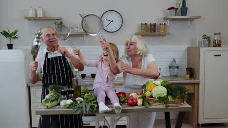 happy vegan senior couple dancing with granddaughter child while cooking vegetables in kitchen