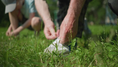 close-up blurred view of an adult tying his shoelace while a little child in the background adjusts her shoe on a grassy field