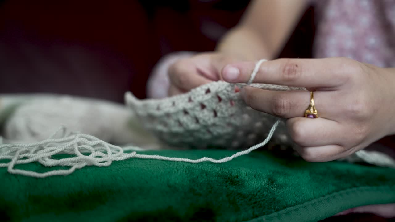 Hands of a woman crocheting with white wool, sitting on a green sofa, creating handmade clothing
