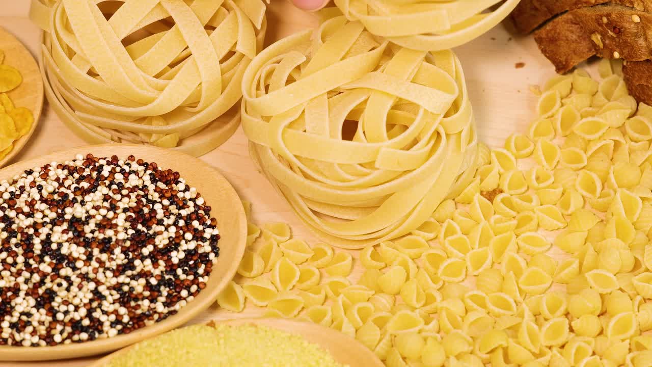 A hand arranges various pasta types and grains on a wooden surface under warm lighting, creating a vibrant, textured display