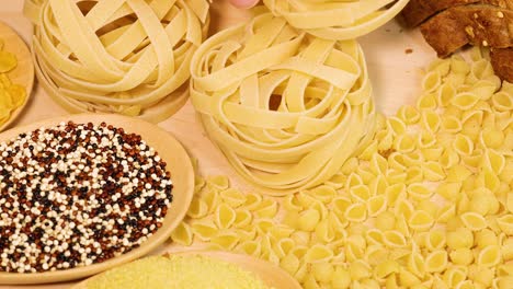 a hand arranges various pasta types and grains on a wooden surface under warm lighting, creating a vibrant, textured display