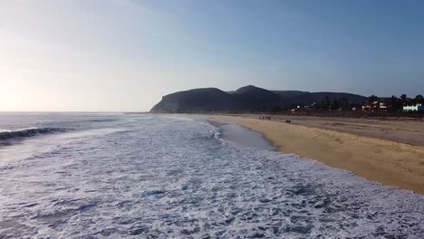 Gente-Con-Perros-En-La-Playa-Mientras-Las-Olas-Rompen-Contra-La-Costa-Arenosa-Durante-La-Puesta-De-Sol.