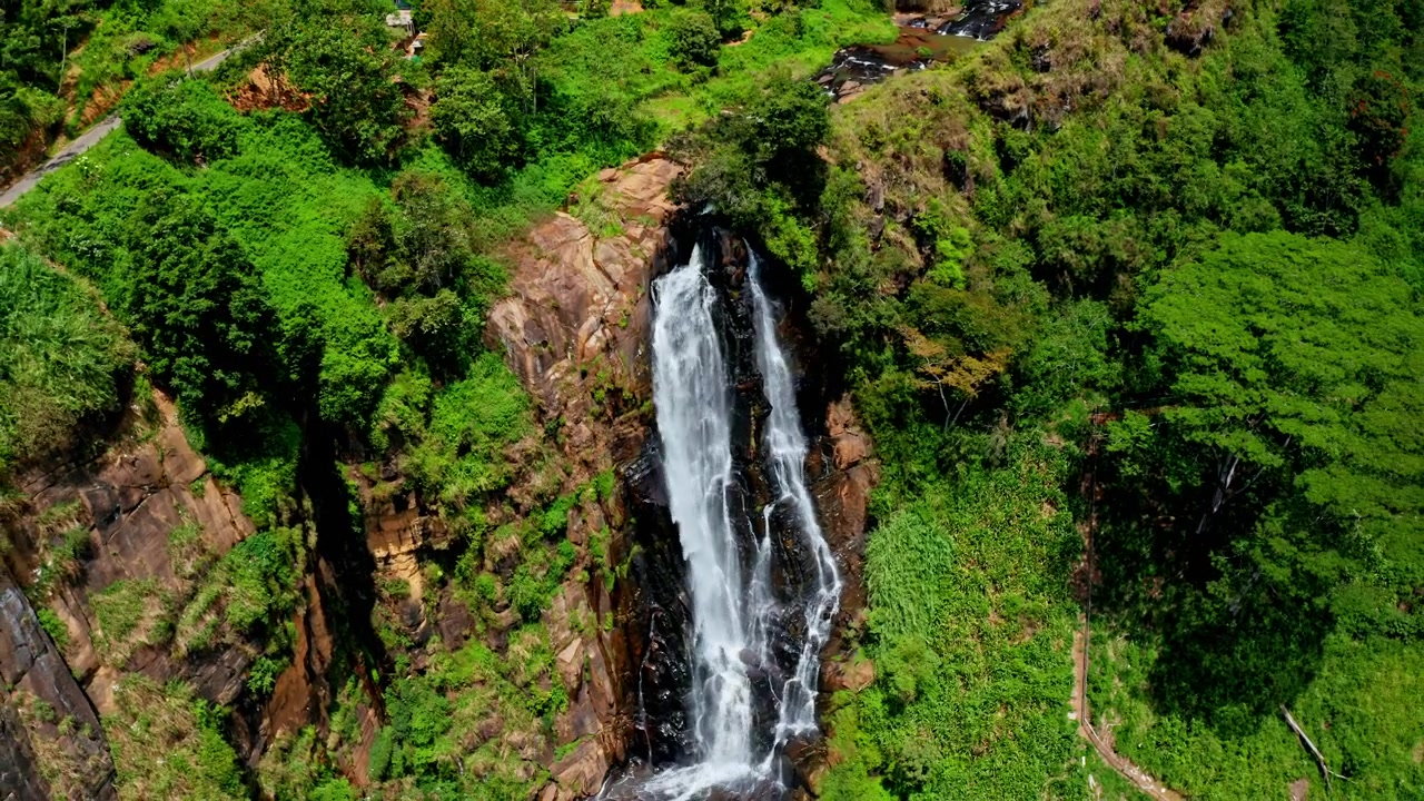 High aerial drone shot of Ella waterfall cascading through tropical forest, rocky cliffs and lush green mountain scenery.