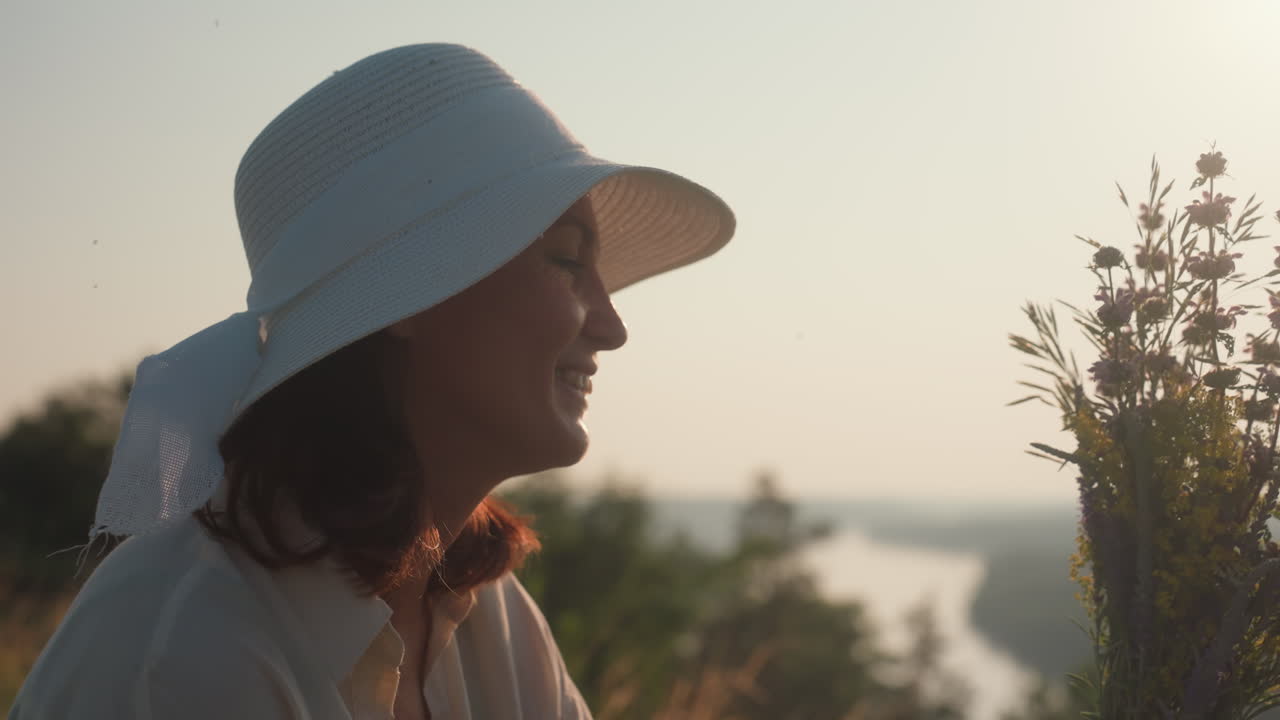 woman in white hat and light shirt holds wildflower bouquet while smiling at it during golden hour, soft sunlight casting glow over grass and trees in background