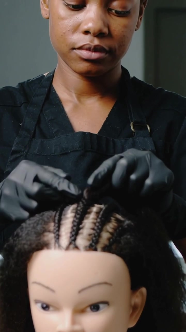 Video verticale: Stilista che divide i capelli e intreccia un manichino in un salone, formando treccine ordinate, con grembiule e guanti.