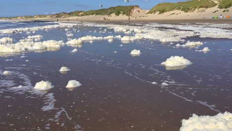 montones de espuma sucia se lanzan por la playa en la playa del mar del norte, mientras que la gente vuela en parapente en el fondo