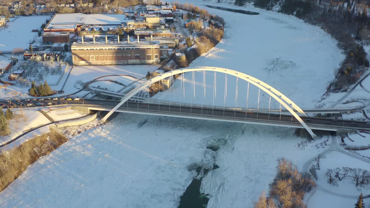 겨울 공중 조감도는 4-5시 화창한 오후 4시 5분에 하얀 현대 월터 데일 다리로 향하는 눈 얼음과 갈라진 찬 물 주머니로 덮인 노스 서스캐처원 강 위를 날아갑니다.