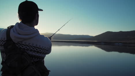 close up silhouette shot of a fisherman casting his lure into a still, glassy, lake in front of gorgeous, snow-capped mountains