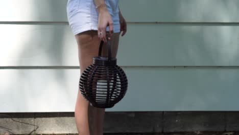 In-the-shade-of-summer-trees,-a-woman-stands-with-a-candle-and-basket-against-the-side-of-a-house