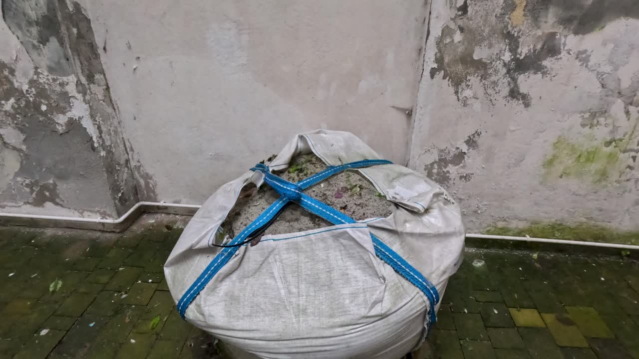 Una grossa borsa coperta con un telo è appoggiata a terra. La borsa è piena di cemento ed è fissata con delle cinghie. L'area presenta pareti usurate e una tonalità verdognola.