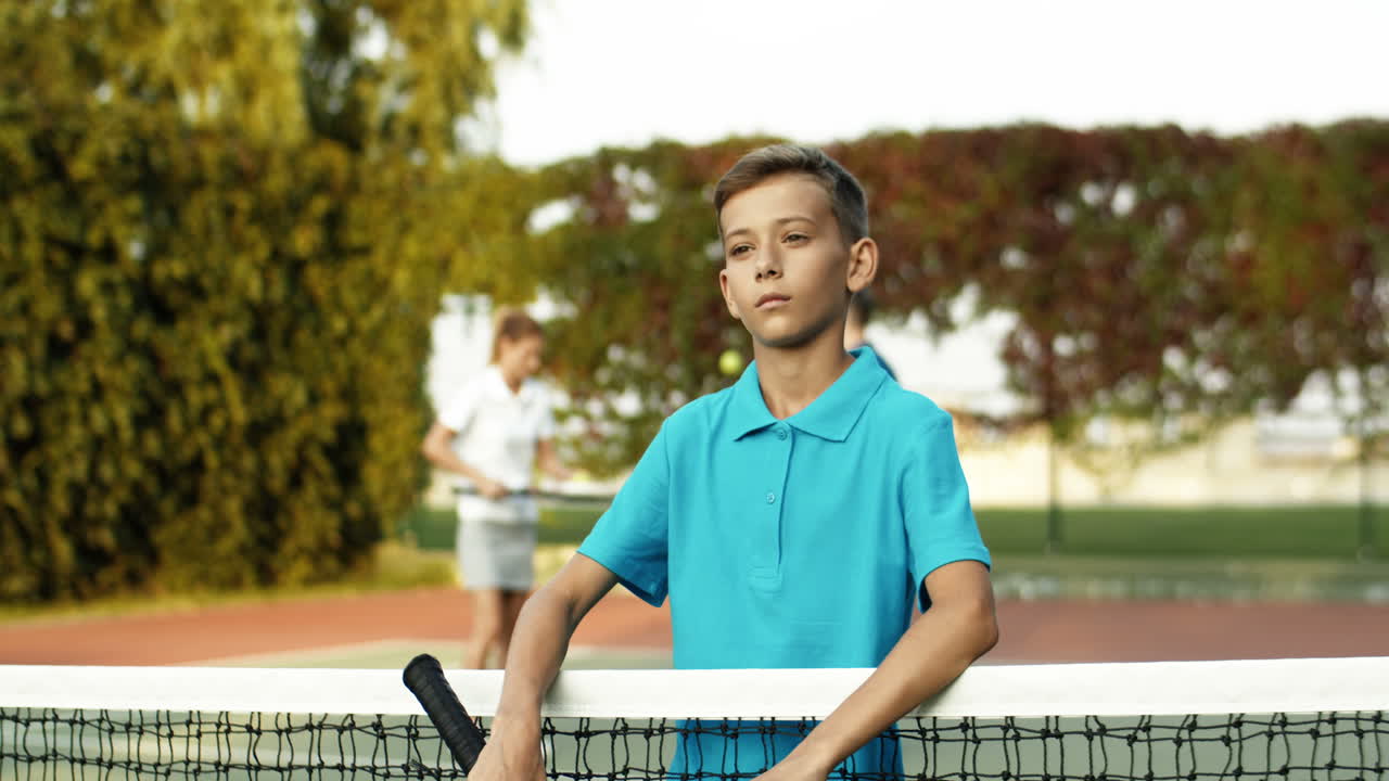 retrato de un lindo adolescente con raqueta de tenis en la mano apoyado en la red y sonriendo alegremente a la cámara