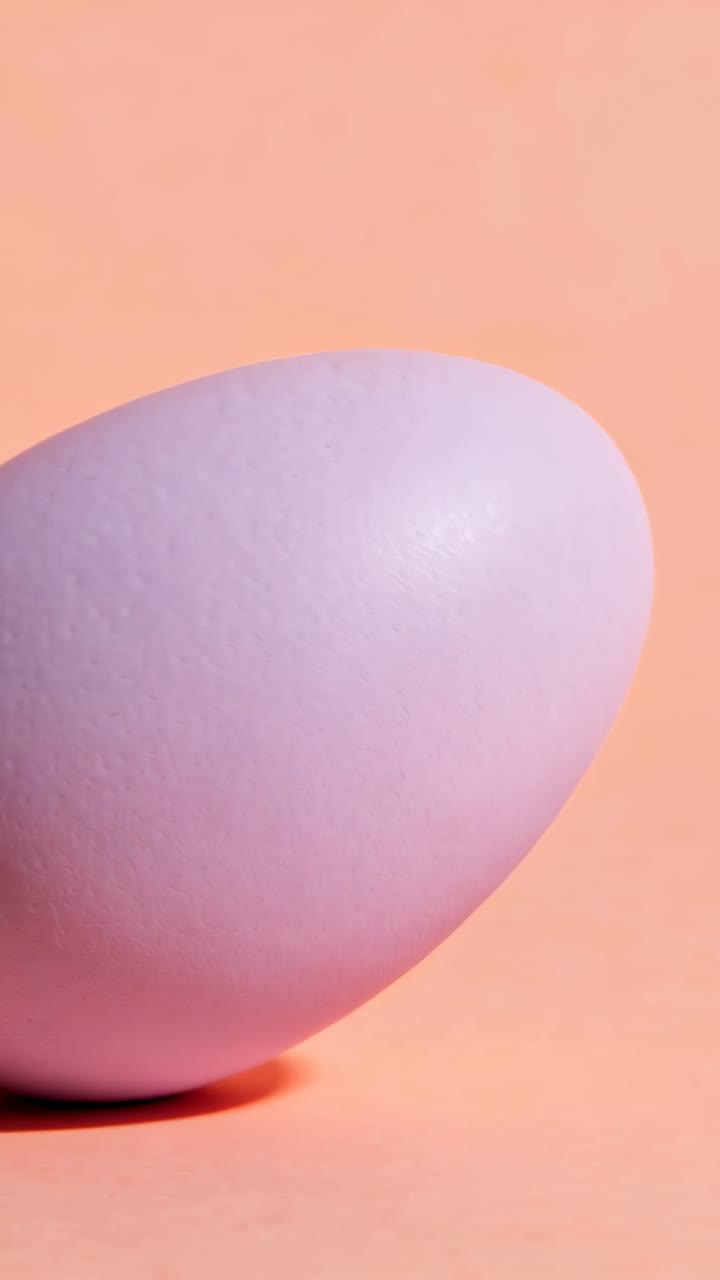 Camera shifting lavender egg tilting, rolling and resting on tip on peach table, highlight, shadow