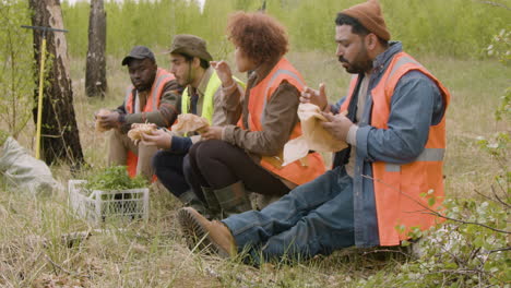 Side-view-of-a-group-of-multiethnic-ecologist-activists-eating-and-talking-in-a-break-sitting-on-a-trunk-in-the-forest