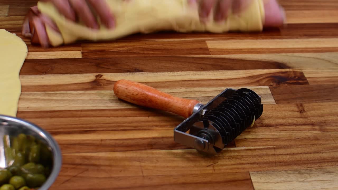 La creación de un patrón decorativo de rejilla en masa de repostería utilizando un rodillo cortador de masa para celosía, lo que refleja una técnica de horneado artesanal y un proceso de preparación de alimentos en una cocina doméstica rústica de madera.