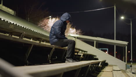 a man wearing a hood is seated alone on bleachers, with a blurred, dimly lit background and an unclear object moving in the distance