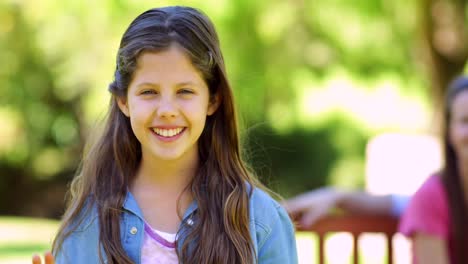 Little-girl-waving-at-camera-with-family-behind-on-park-bench