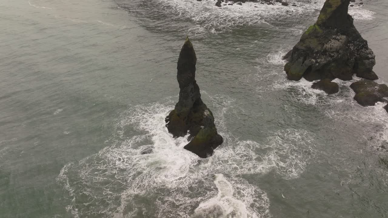 Aerial view of a towering Reynisdrangar sea stack emerging from the ocean near Reynisfjara beach, Iceland, with waves crashing against the volcanic formation.