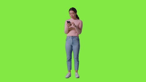 full length studio shot of woman scrolling through messages or content on mobile phone in front of green screen