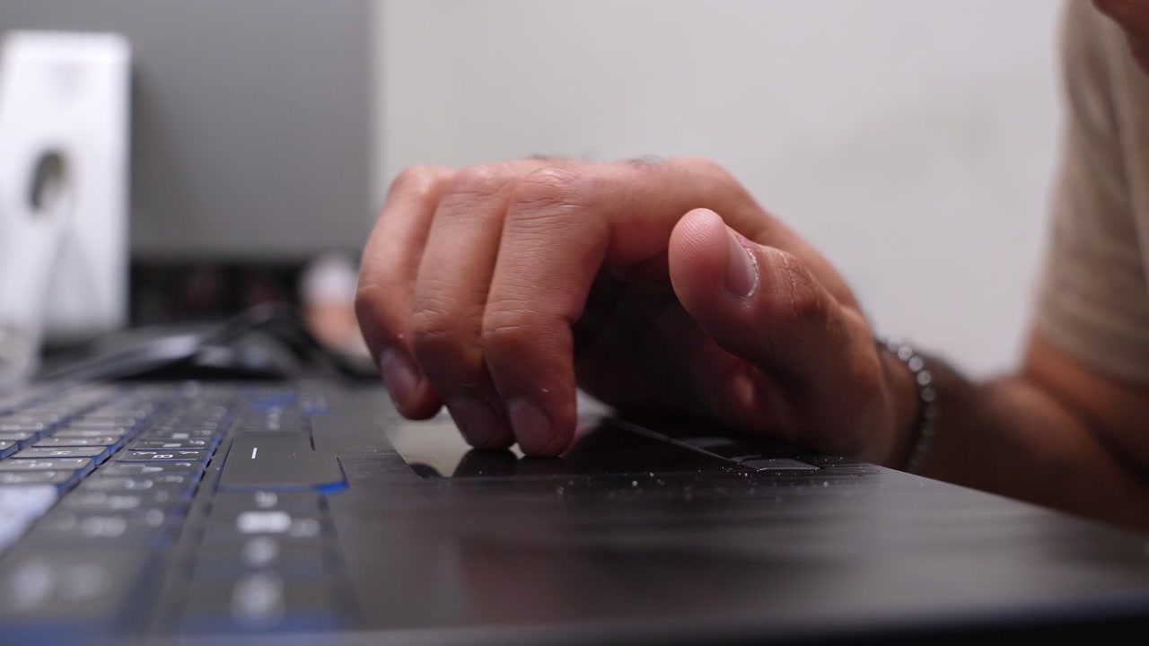 En mands hånd navigerer en bærbar computers trackpad på et skrivebord i et moderne hjemmekontor i dagslys. Cinematisk optagelse med lav dybdeskarphed, ideel til reklamer for virksomheder og teknologiske livsstilsprodukter.