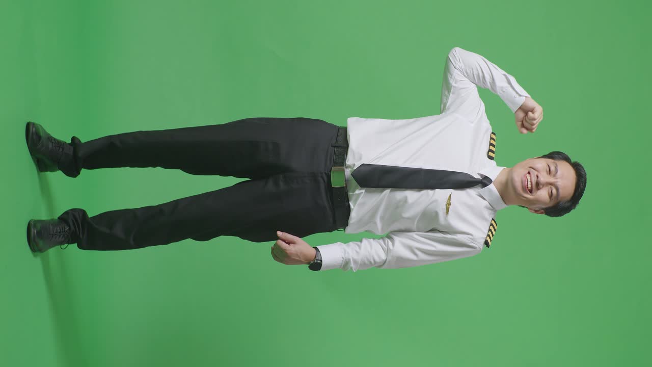 녹색 스크린 배경 스튜디오에 서 있는 동안 미소 짓고 팔다리를 구부리는 아시아 남성 조종사의 몸이 가득 차 있습니다.