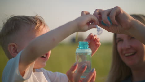 una mujer alegre con un vestido azul abre una pequeña botella de espuma en un campo cubierto de hierba mientras un joven feliz con una camiseta blanca observa con deleite, tomando con entusiasmo la varita y soplando burbujas en el aire