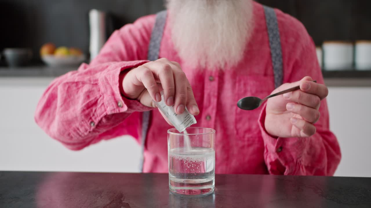 retrato en primer plano de un hombre con cabello gris y una barba exuberante en una camisa rosa vierte medicina blanca en un vaso transparente de agua para sí mismo y lo agita en un apartamento moderno