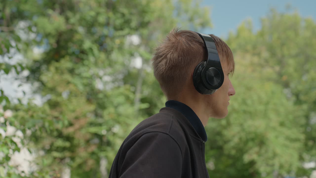 Hvit mann med hodetelefoner utendørs som lytter til musikk mens han spaserer gjennom en grønn park, nærbilder i profil fanger opp tankefulle uttrykk, avslappet genser, lett bris, solbelyst løvverk, subtilt