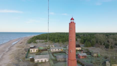 Vista-Aérea-Del-Faro-Akmenrags-De-Color-Rojo,-Costa-Del-Mar-Báltico,-Letonia,-Playa-De-Arena-Blanca,-Mar-En-Calma,-Día-Soleado-Con-Nubes,-Amplia-Toma-Ascendente-De-Drones-Avanzando