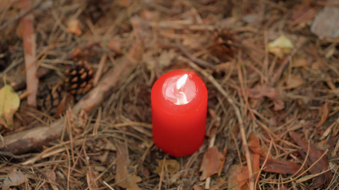 Close de uma única vela vermelha queimando continuamente no chão da floresta entre folhas secas e pinhas, com uma chama quente tremeluzindo suavemente sob a luz natural, simbolizando a meditação e a energia espiritual sagrada