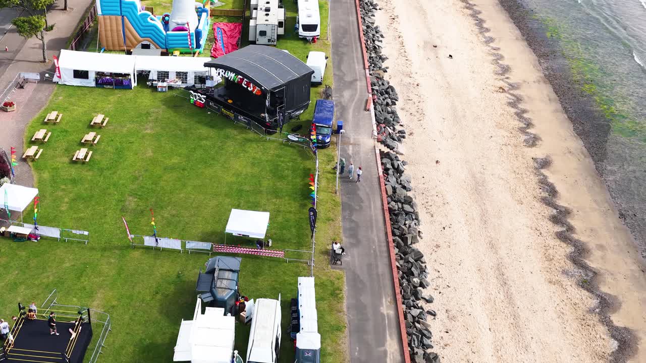 Drone glides above seaside fairground, amusement rides, tents, and beach under bright daylight conditions