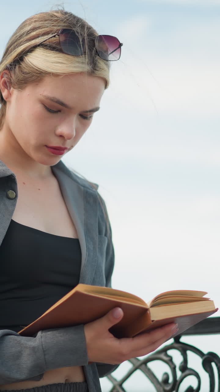 Lady in open grey shirt holding an open book with right hand on pages and left hand beneath, reading with focus, wind softly blows the book's pages, blurred background shows water body
