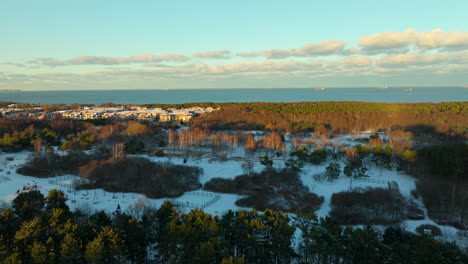 ascending drone shot of forest landscape with snow and gdansk district in background at sunrise - blue baltic sea in backdrop