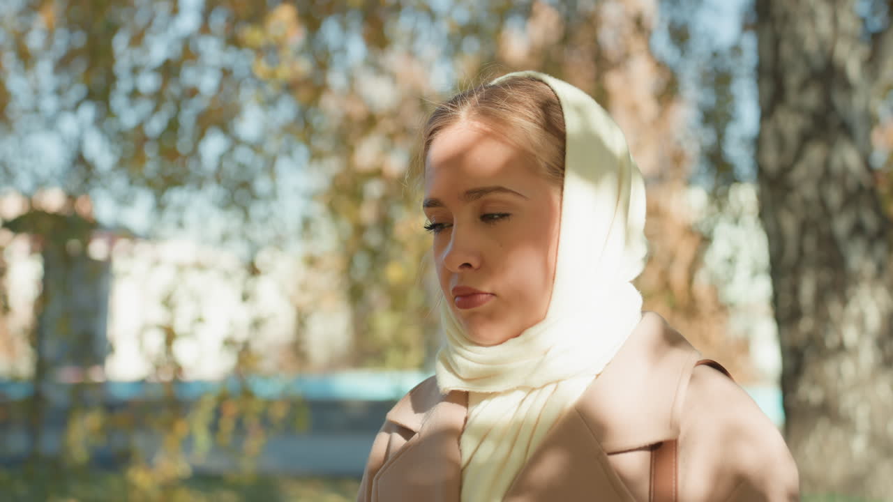 Huş ağacının altında nehir kenarında, yüzü krem rengi eşarp ve bej paltoyla çerçevelenmiş Kafkasyalı kadın, aşağıya doğru bakış, sarı yaprakların arasından süzülen güneş benekli gölgeler, düşünceli ruh hali, sinematik profil çekimi, şehir sahili.