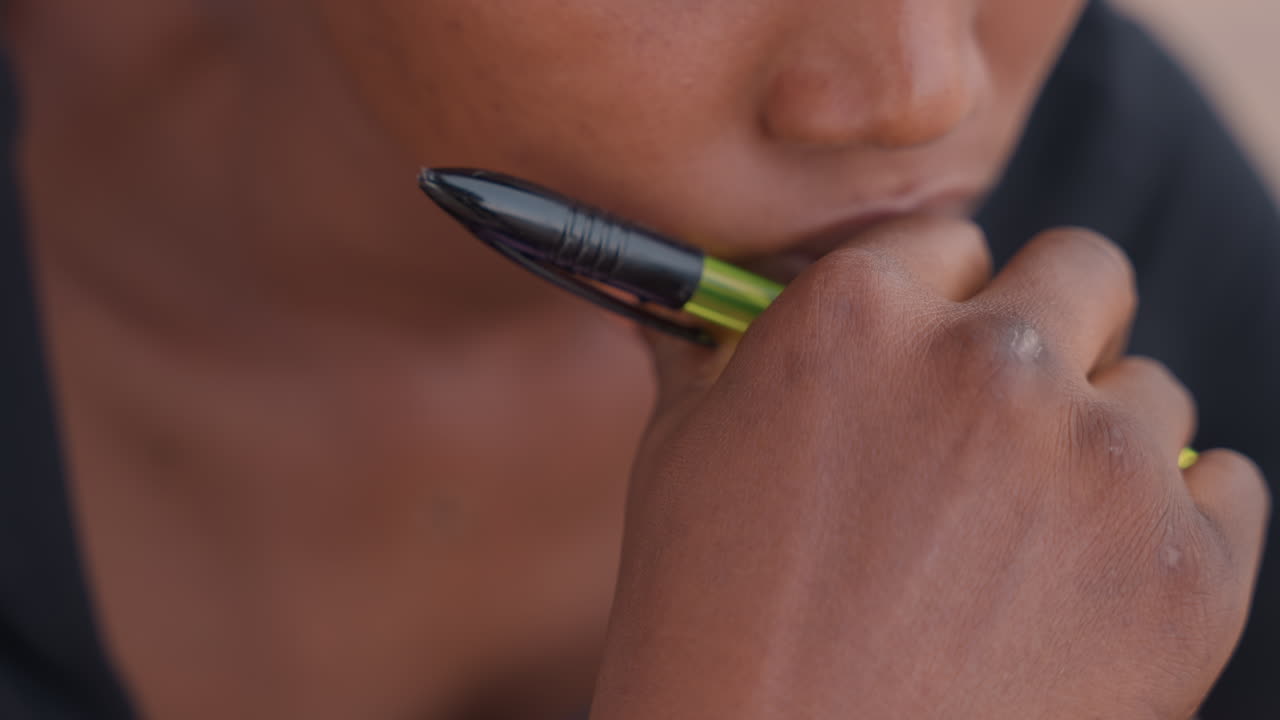 Primer plano de mujer pensativa sosteniendo un bolígrafo, labios cerca de este y mano en la barbilla, luz suave y poca profundidad de campo, ambiente de estudio íntimo, planificando notas y con vacilación creativa, textura sutil de la piel.